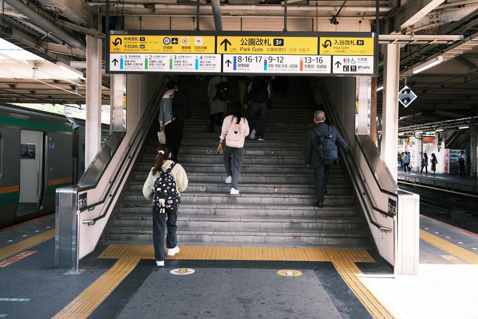 The image depicts a busy train station entrance with a wide set of grey concrete stairs leading up to a platform, framed by stainless steel handrails on each side. Several commuters are ascending the